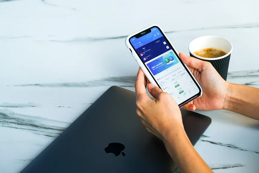 Hände halten ein Smartphone mit geöffneter Krypto-App, daneben ein Kaffeebecher und ein Laptop