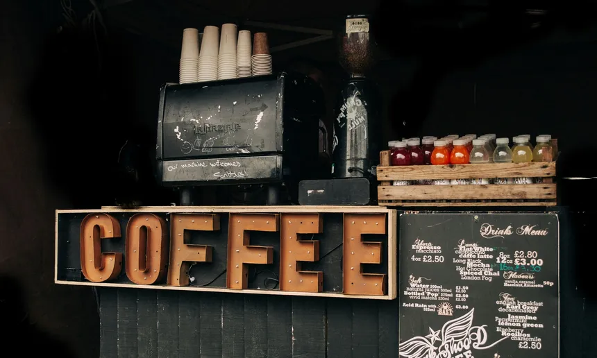 Dunkler Kaffeestand: rostbraune Leuchtbuchstaben „COFFEE“ in einem Holzrahmen, darüber eine abgenutzte Espressomaschine mit gestapelten Pappbechern und eine Kaffeemühle, rechts eine Kiste mit bunten Saftflaschen und eine Kreidetafel mit der Getränkekarte